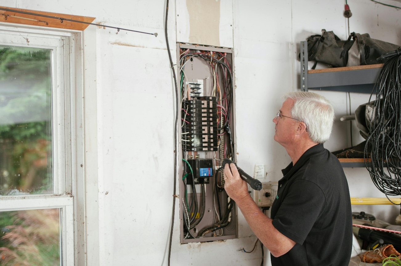 A homeowner inspecting an open residential electrical panel with a flashlight